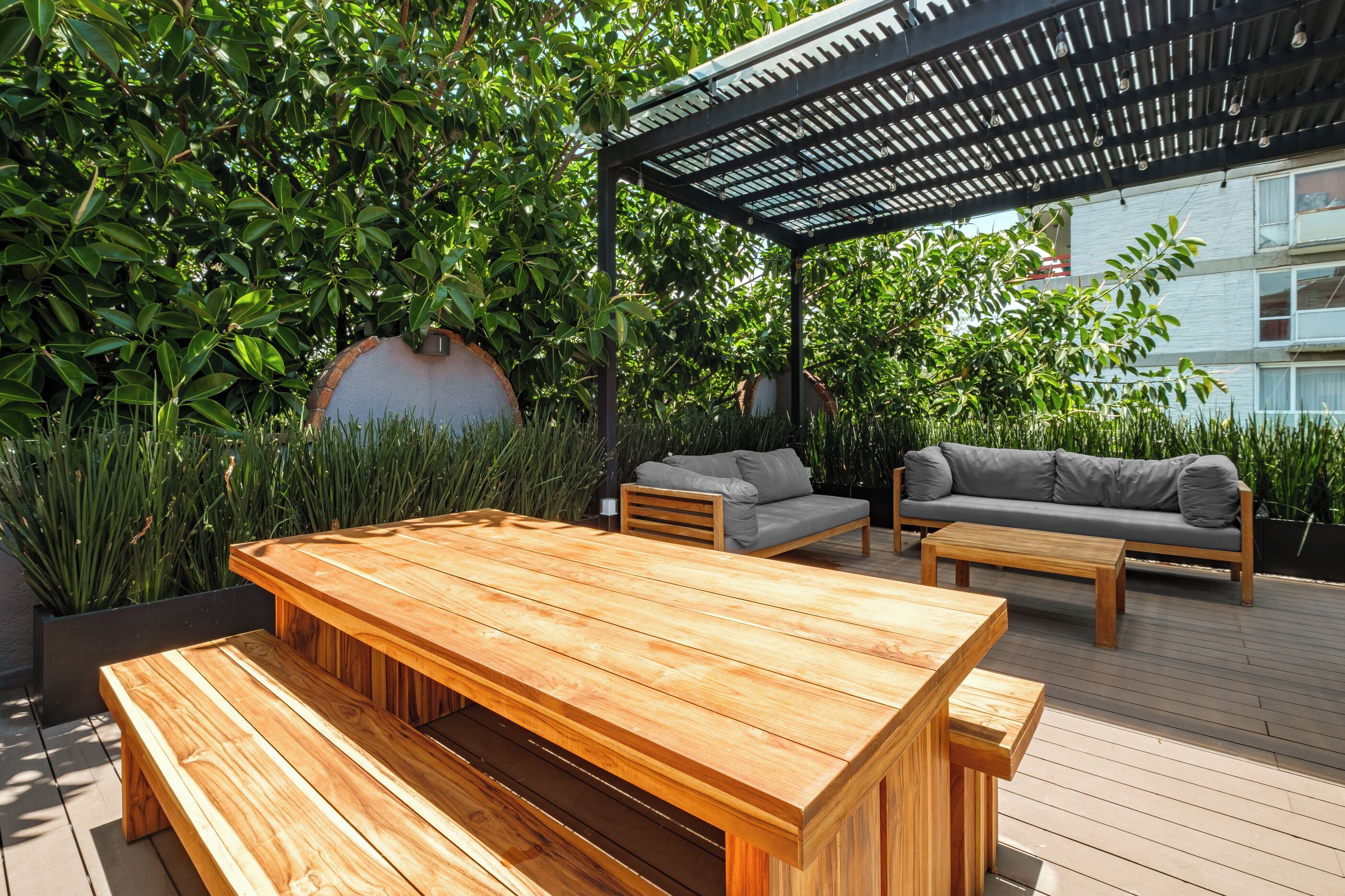 Une terrasse en bois avec une grande table et des bancs, entourée d’une végétation luxuriante et dotée d’un espace salon abrité.