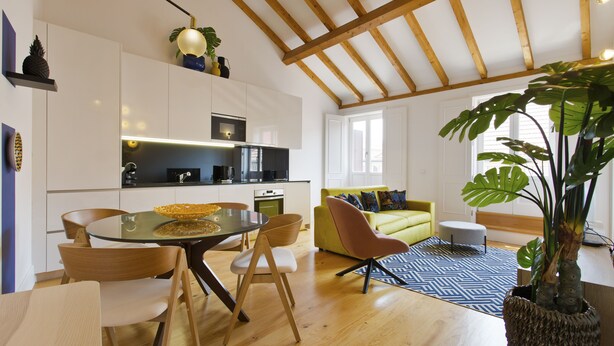 A modern kitchen with white cabinets, a dining table with chairs, a yellow sofa, and a large plant. - Boutique Rentals- The Alma Apt w/balcony (Porto)