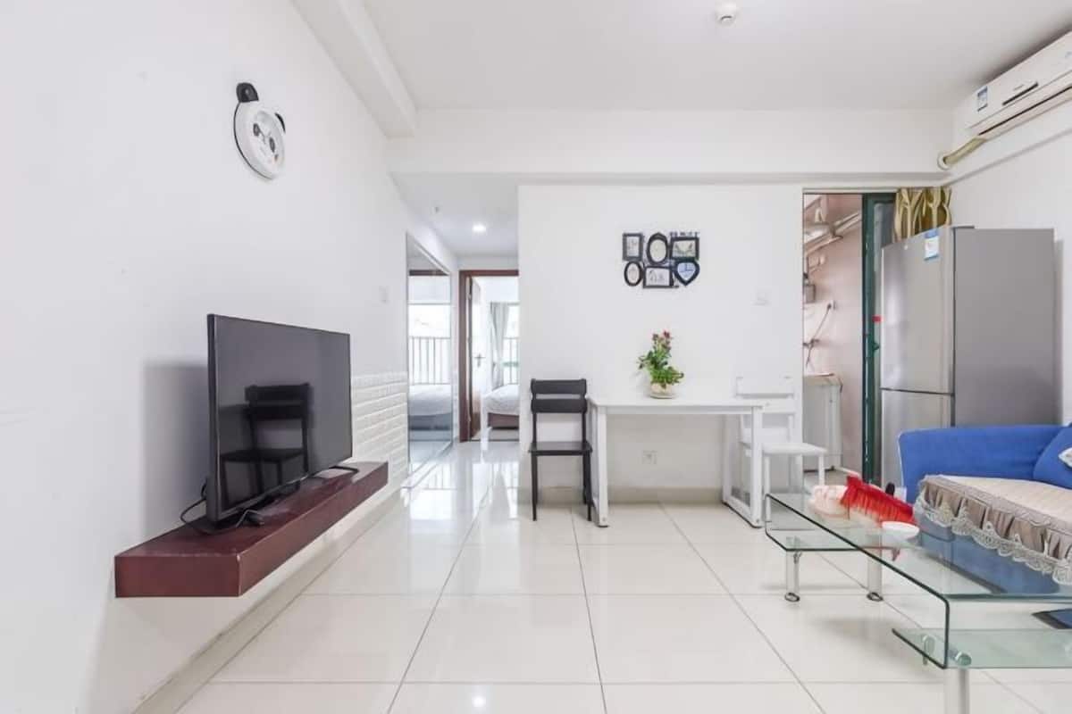 Una sala de estar moderna con un televisor de pantalla plana, una mesa de centro de vidrio, un sofá y un comedor con una mesa y sillas blancas.