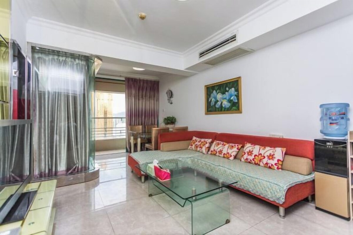Uma sala de estar com uma mesa de centro de vidro, um sofá vermelho com almofadas florais, um dispensador de água e um quadro na parede.