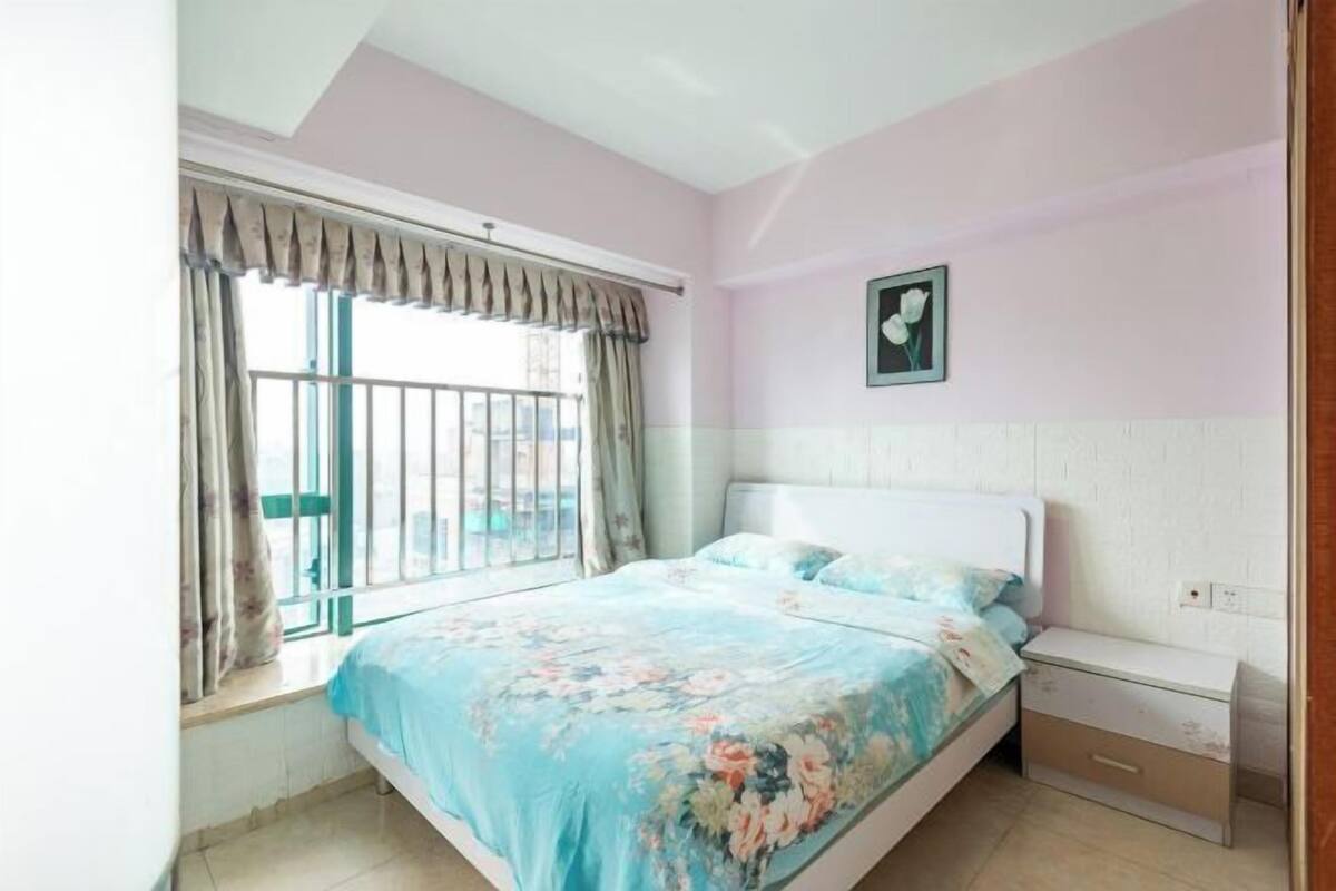 Un dormitorio con cama, mesita de noche, ventana con cortinas e imagen colgada en la pared.