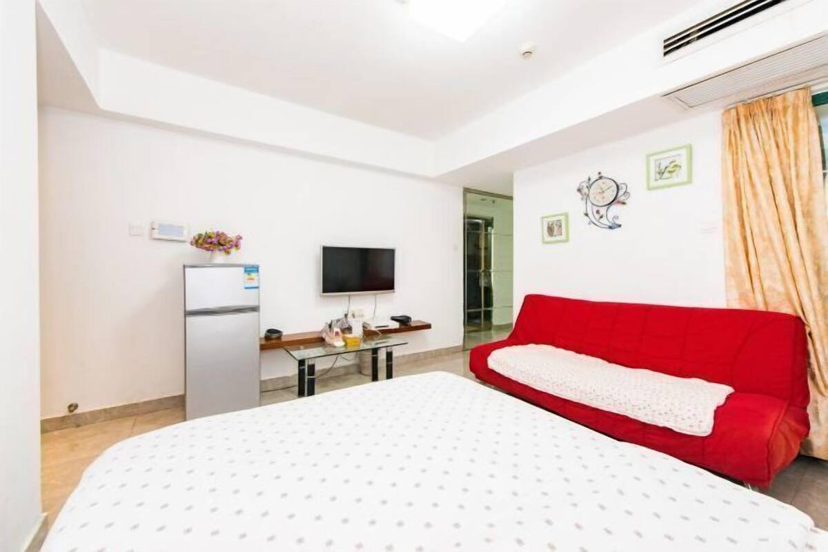 Habitación de hotel con cama, sofá rojo, televisión, refrigerador y ventana con cortinas.