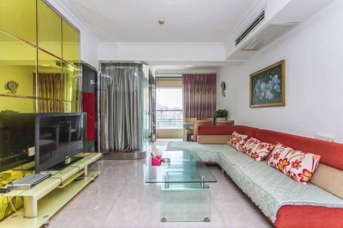 Una sala de estar con un sofá, una mesa de centro de vidrio, un televisor sobre un mueble bajo y un ventanal con cortinas.