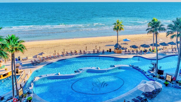 A pool with a circular design, surrounded by lounge chairs and umbrellas, with a beach and ocean in the background. - Oceanfront View- Sandy Beach-Sonoran Sea Resort (Puerto Peñasco)