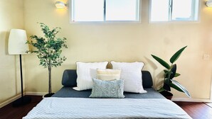 Un dormitorio con cama, dos ventanas, una lámpara de pie y una planta en maceta.