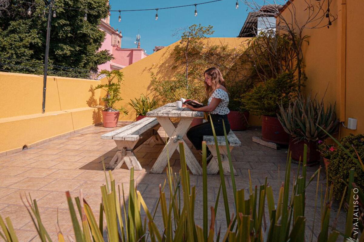 A woman reading at a wooden table in a courtyard with potted plants, a yellow wall, and a tower in the background.
