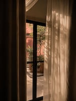 Janela com cortinas leves, vista de um pátio com plantas e um edifício ao fundo.