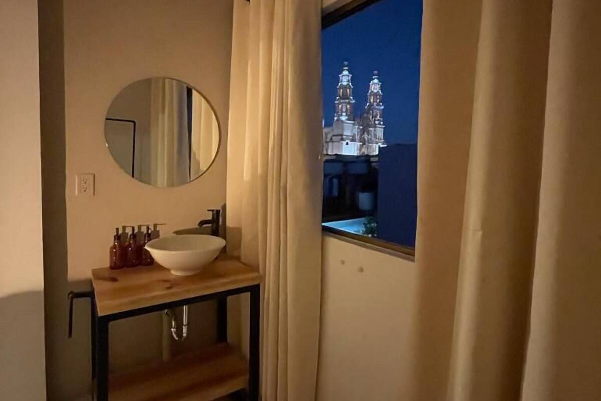 A bathroom with a round mirror, a sink, and a window with curtains.