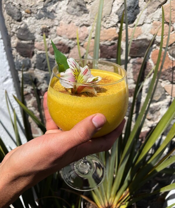 A hand holding a glass with a yellow beverage, garnished with a flower, against a stone wall background. - Hotel Boutique Casa Damiana (Lagos de Moreno)