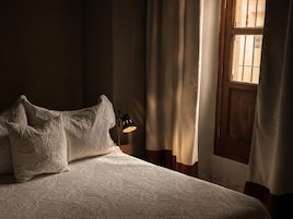 Cama bem arrumada com colcha estampada e duas almofadas, abajur e janela com cortinas.