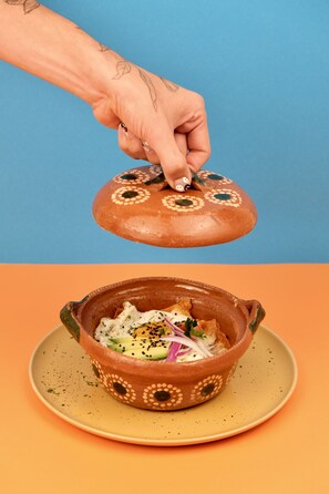 A hand lifting a lid from a clay pot with a floral design, revealing a dish with rice, vegetables, and a dollop of sauce on a plate. - Hotel Boutique Casa Damiana (Lagos de Moreno)
