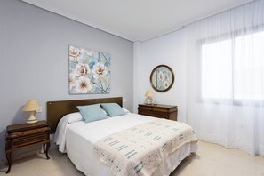 A bedroom with a bed, two nightstands, a wall-mounted mirror, and a large window with sheer curtains.
