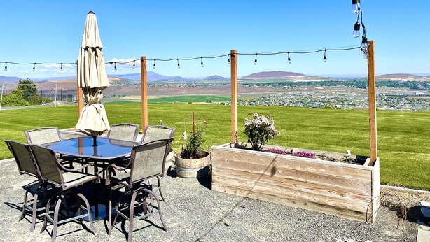 An outdoor patio with a table and chairs, string lights, and a view of a town and hills. - Wine Country Getaway with Amazing Views (Kennewick)