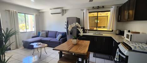 A modern living room with a sofa, coffee table, and a dining area with a wooden table and chairs. There is a kitchen with a microwave, sink, and cabinets.