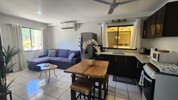 A modern living room with a sofa, coffee table, and a dining area with a wooden table and chairs. There is a kitchen with a microwave, sink, and cabinets.