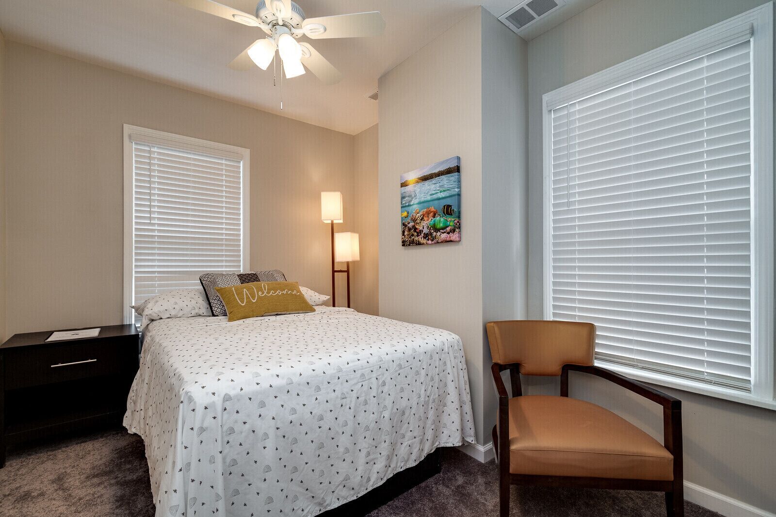 Une chambre à coucher avec un lit, une table de chevet, une chaise, un ventilateur de plafond et un tableau accroché au mur.