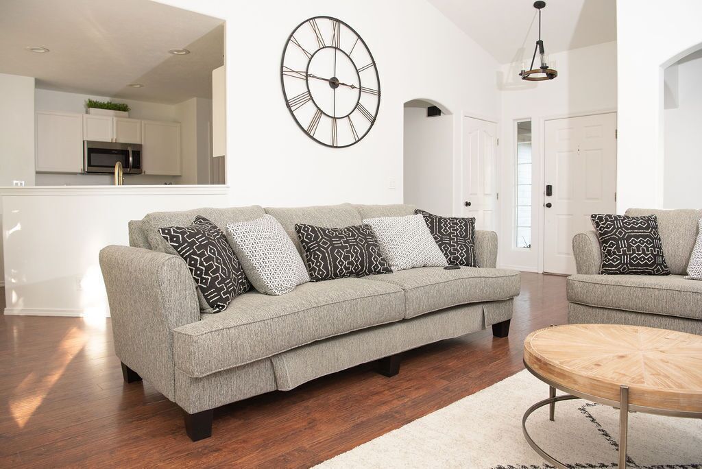 A modern living room with a grey sofa, patterned cushions, a round wooden coffee table, and a large wall clock.