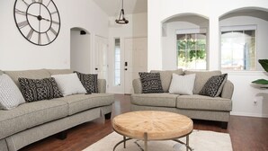 Un salon avec deux canapés gris, des coussins à motifs, une table basse ronde en bois et une grande horloge murale.