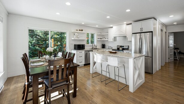 A modern kitchen with white cabinetry, a central island, and a dining area with wooden chairs. - Peaceful Parkside Cottage (Courtenay)