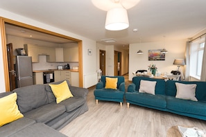 A modern living room with a grey sofa and teal armchairs, a kitchen in the background, and a dining area with a table and chairs.