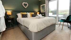 A bedroom with a large bed, a heart-shaped wall decoration, a nightstand with a lamp, and a view of the outdoors through a sliding door. - Beths Bothy -  a pod that sleeps 2 guests  in 1 bedroom (Gatehouse of Fleet)