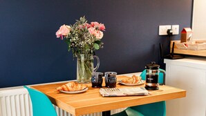 A dining area with a wooden table, two teal chairs, a vase with flowers, a coffee maker, a croissant, and a newspaper.