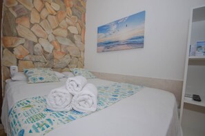 Un lit bien fait, recouvert d’une couvre-lit à motifs, un mur en pierre, un tableau représentant une scène de plage et une étagère avec une télécommande.