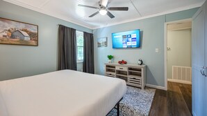 Une chambre à coucher avec un lit, un ventilateur de plafond, un téléviseur fixé au mur, une petite table avec une plante et une voiture jouet, et une fenêtre avec des rideaux.