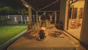 An outdoor dining area with a table set for a meal, surrounded by a covered patio with a view of the garden and a building entrance. - Vila Gameleira Garça Torta (Maceió)