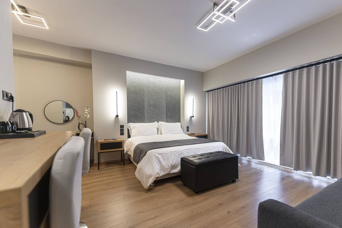 A modern hotel room with a large bed, a desk, a chair, a small table with a kettle, and a large window with curtains.