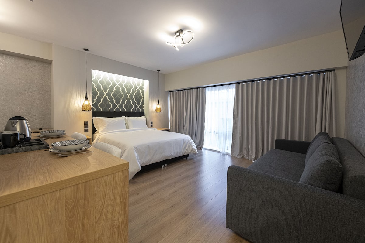 A modern hotel room with a wooden desk, a bed, a sofa, and a large window with curtains.