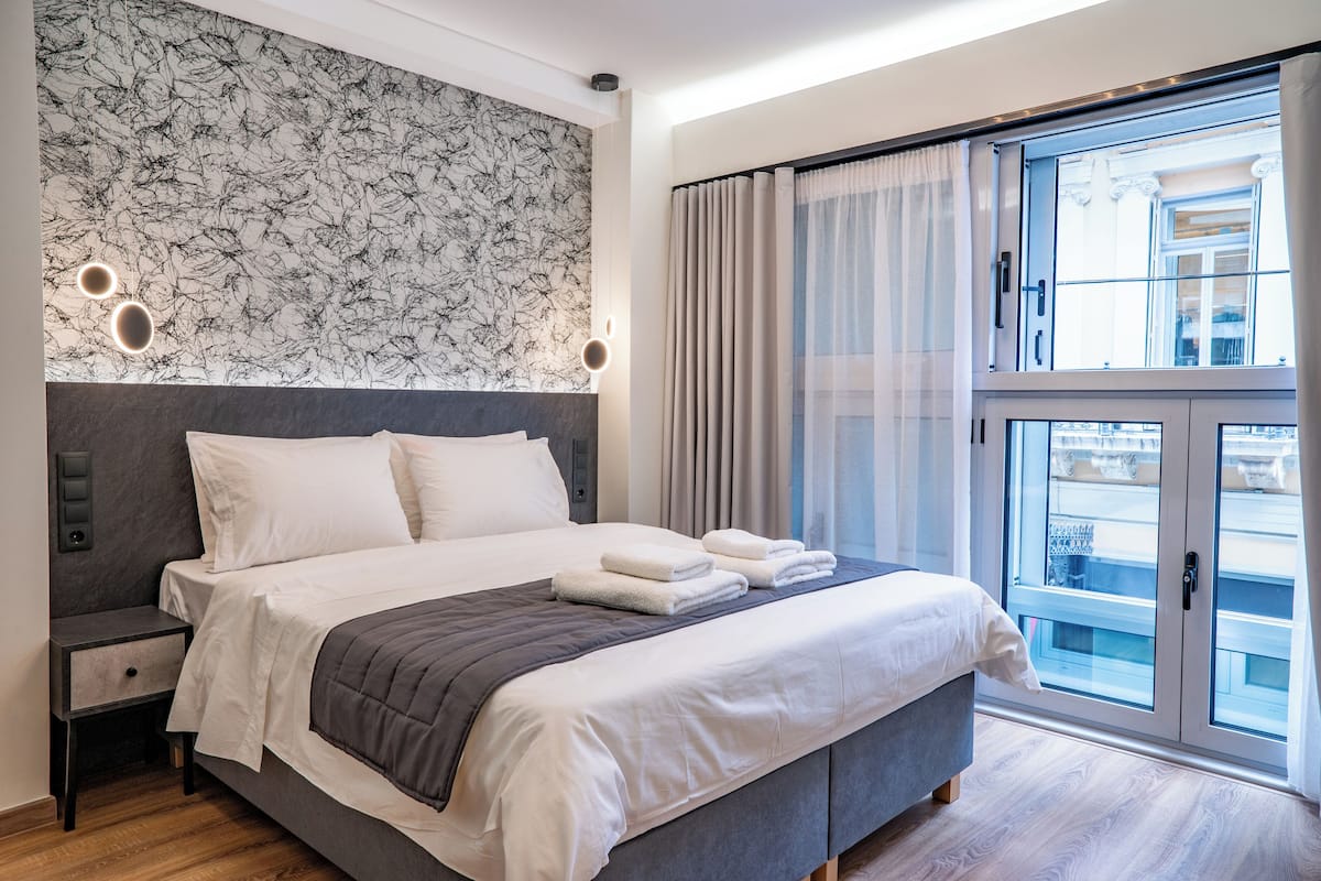 A modern hotel room with a large bed, a headboard with a patterned wallpaper, a bedside table, and a window with curtains.