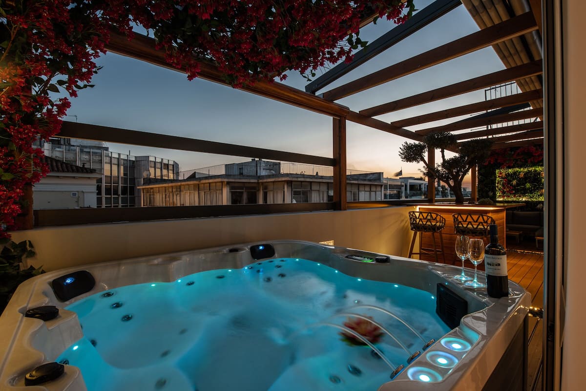 A hot tub with a view of buildings, a bar area with a bottle of wine, and a wooden pergola with flowers.