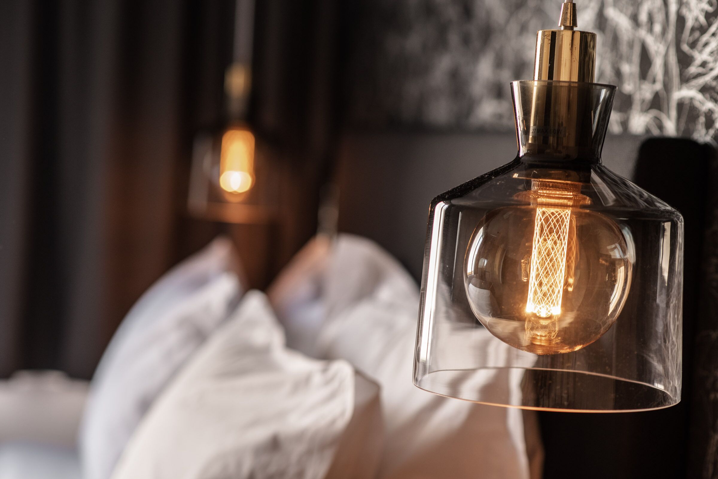 Un luminaire suspendu en forme de bouteille en verre transparent, avec une ampoule à filament visible à l’intérieur, placé dans une pièce faiblement éclairée.