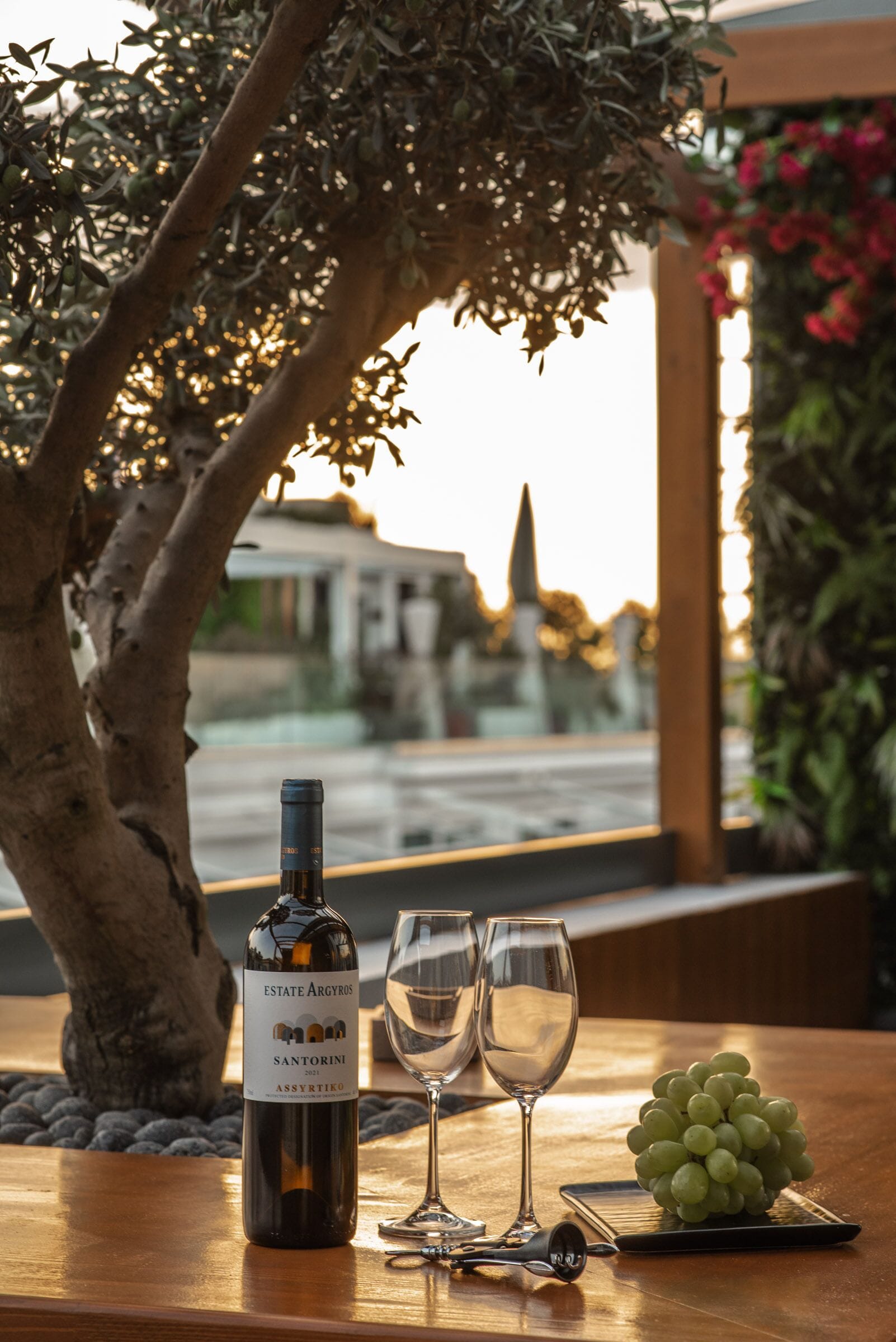Una botella de vino, dos copas y un racimo de uvas sobre una mesa con vistas a un paisaje urbano y un árbol.