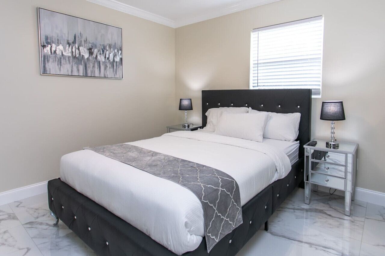 Une chambre à coucher avec un lit, deux tables de chevet avec des lampes, une fenêtre avec des stores et un tableau au mur.
