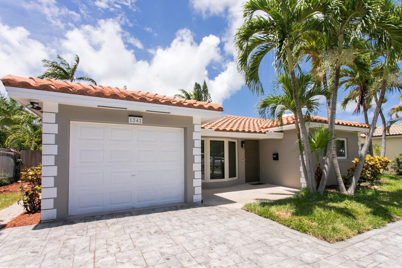 A single-story house with a white garage door, a red-tiled roof, and a small front yard with palm trees.