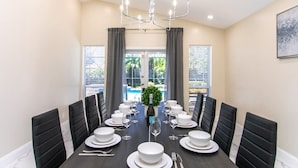 Ein Esszimmer mit einem langen Tisch, der für eine Mahlzeit gedeckt ist, schwarzen, gepolsterten Stühlen, einer Pendelleuchte und Blick ins Freie durch große Fenster.