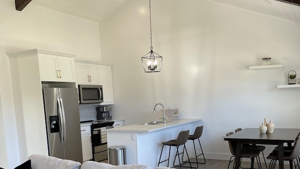 A modern kitchen with white cabinets, a stainless steel refrigerator, and a microwave. There is a dining area with a table and chairs, and a sofa with cushions.