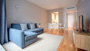 A modern living room with a grey sofa, a dining area with a white table and chairs, and a TV on a wooden stand.
