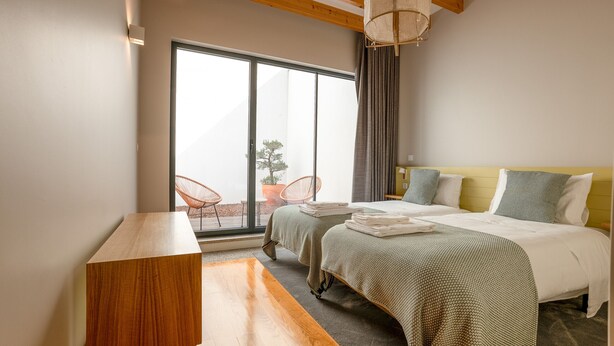 A hotel room with two beds, a wooden side table, a large window with a view of the outdoors, and a ceiling with exposed beams.