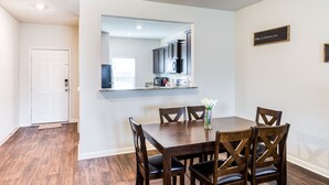 Een eetgedeelte met een houten tafel en stoelen, een vaas met bloemen, en een keuken die in de spiegel te zien is.