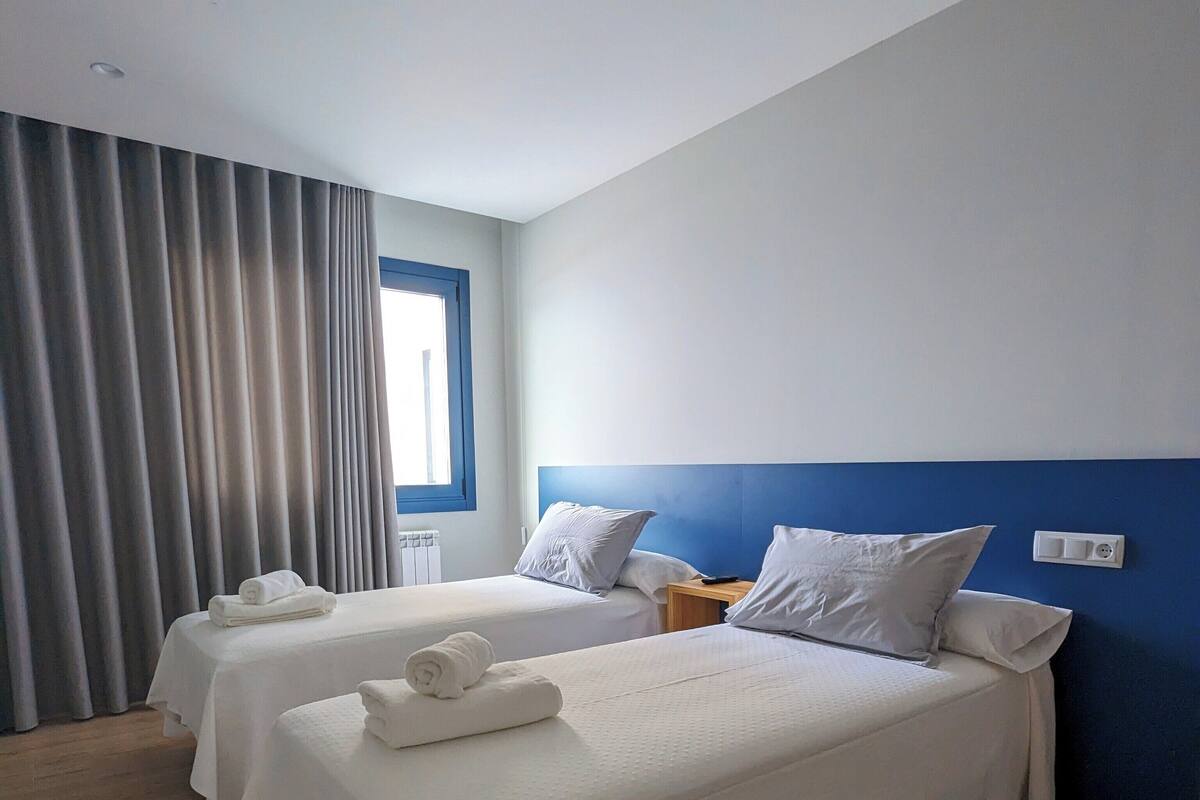 A hotel room with two beds, a window with curtains, a bedside table, and a wall switch.
