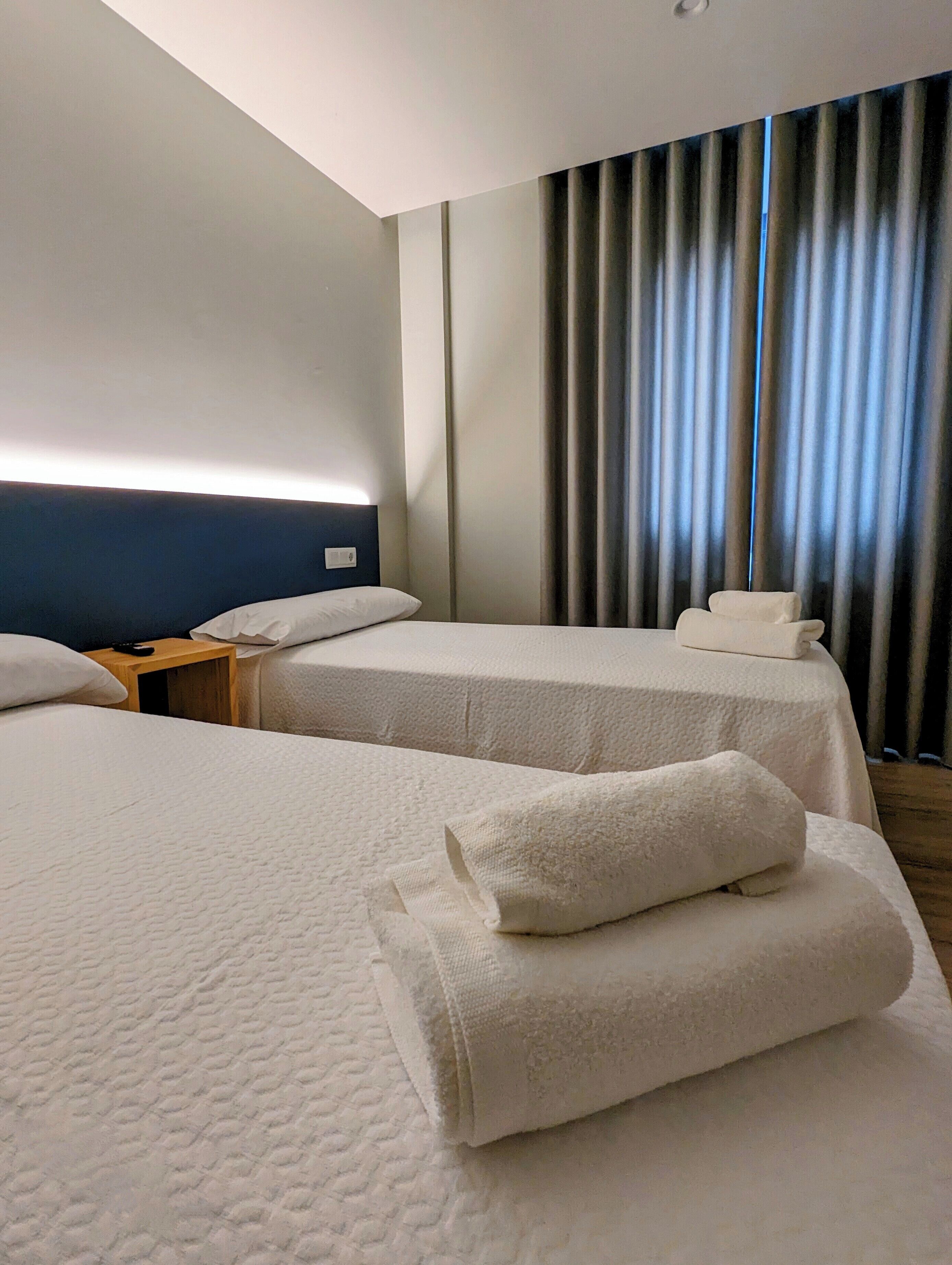 A hotel room with two beds, a nightstand, a headboard, and a view of the outside through curtains.