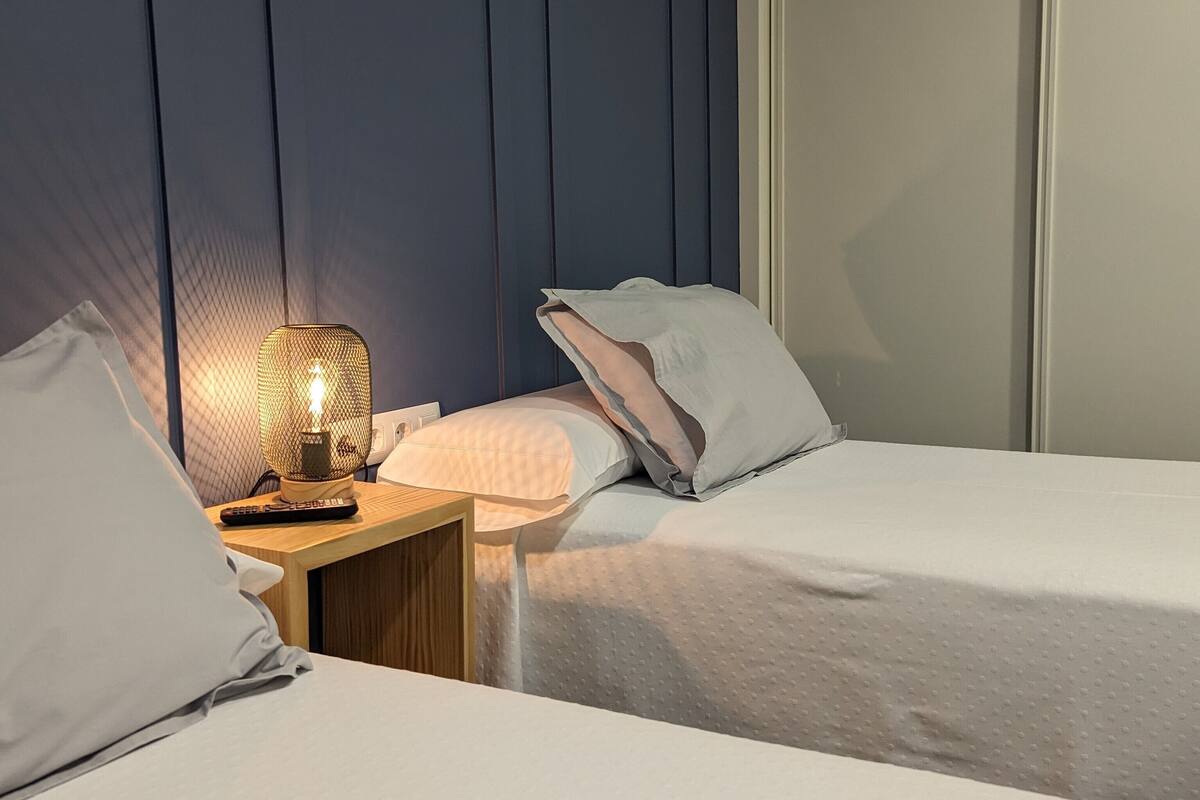 A bedroom with two beds, a nightstand with a lamp, and a window with blinds.