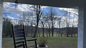 A porch with a rocking chair and a potted plant, overlooking a grassy yard with trees and a distant building. - Beautiful quiet retreat near Oneida, Big South Fork, and ATV trails! (Oneida)