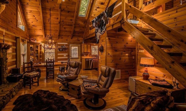 A log cabin interior with a cozy fireplace, leather chairs, a dining area, and a staircase.