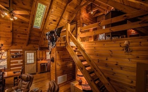 A rustic cabin interior with wooden walls, a staircase, a dining area, and a kitchen.