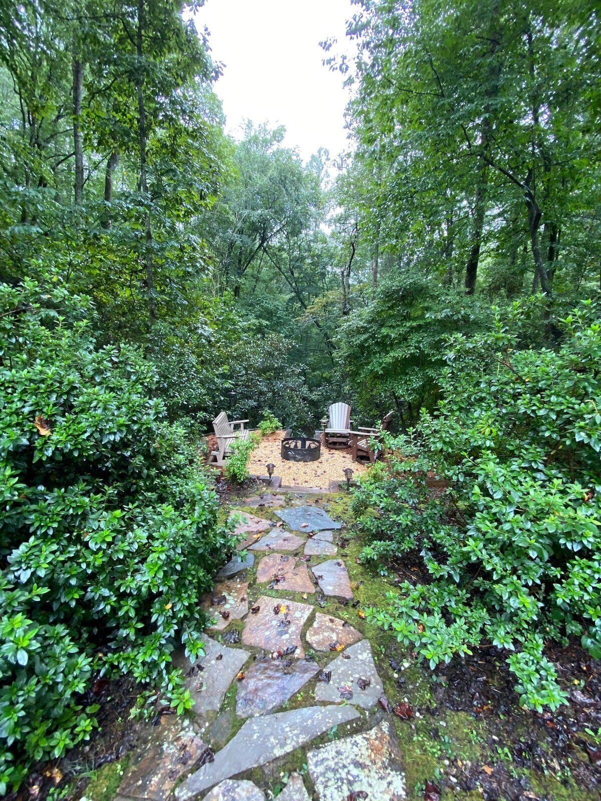 A stone pathway leading through a lush forested area with a fire pit and wooden chairs.