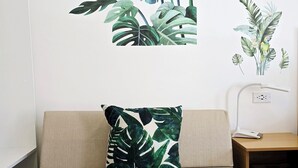 A beige sofa with a tropical leaf-patterned pillow, a white lamp, and a wall decal with a bird and leaves. - Habitación Independiente #2 , Entre Airport y SJ, Privada (Uruca)